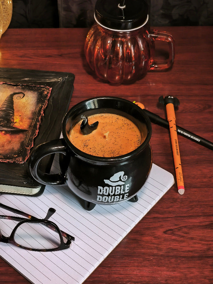 Photo d'une bougie orangée dans une tasse en forme de chaudron noir, et dans laquelle est posée un pendentif en croissant de lune en obsidienne noire.